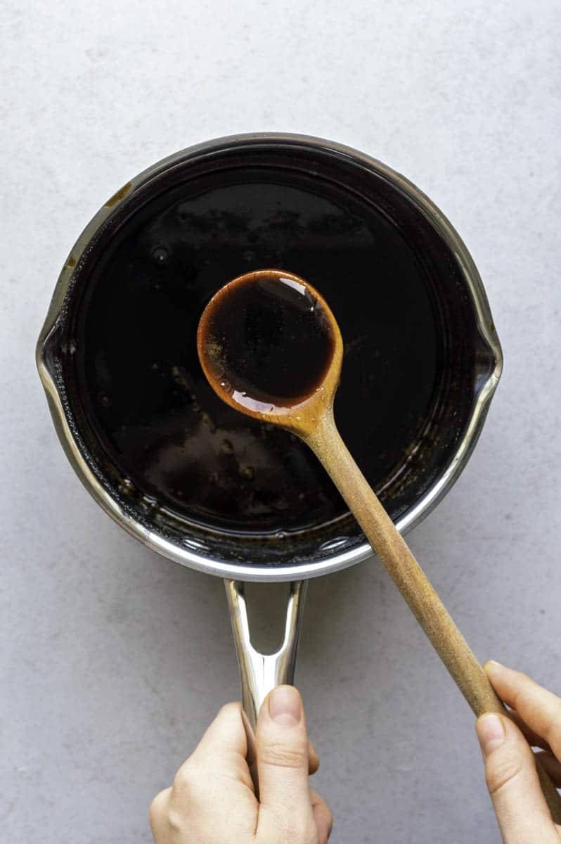 Brown sugar syrup in a stainless-steel saucepan. A wooden spoon filled with syrup hovering on top of the saucepan.