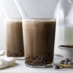 Two glasses of chocolate boba with glass straws on a white marbled board.