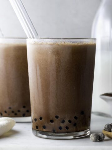 Two glasses of chocolate boba with glass straws on a white marbled board.