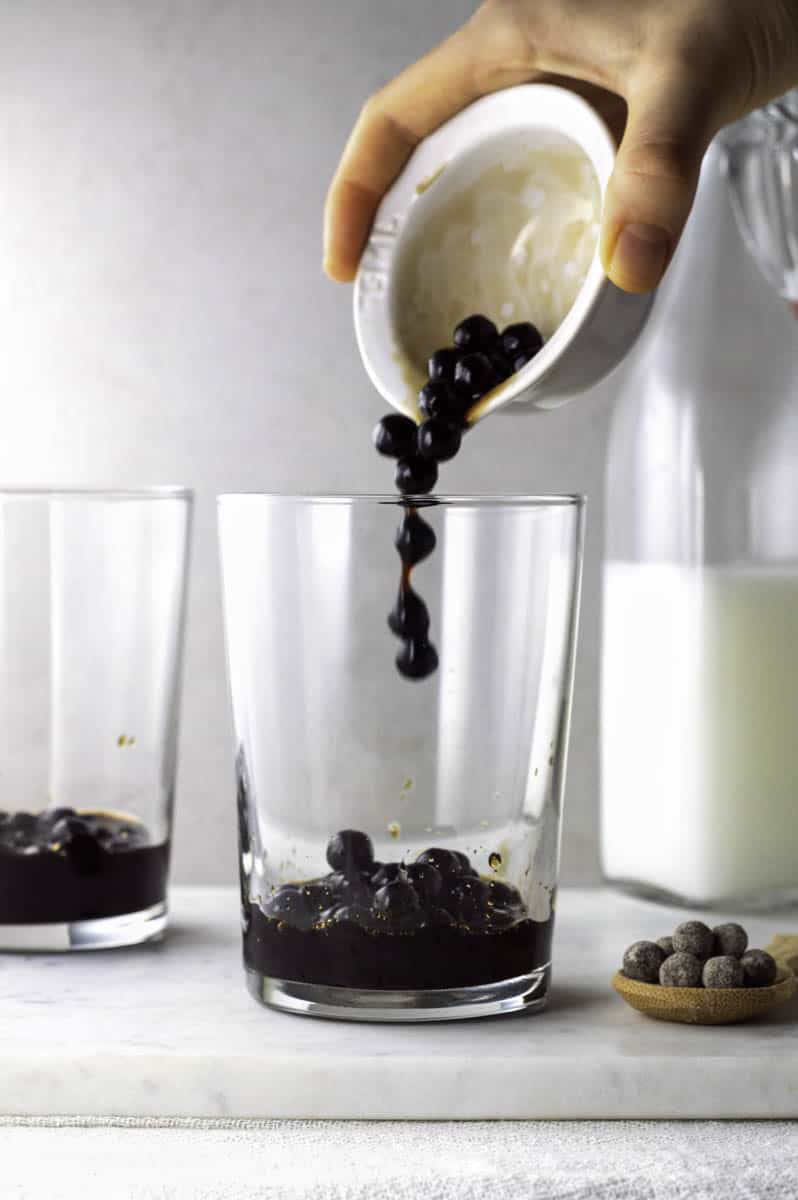 A hand adding pouring tapioca pearls from a small white bowl to a large glass.