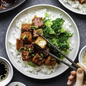 Gochujang tofu served with steamed broccoli over a bowl of rice. A hand picking up a piece of tofu with a bite take off of it with dark brown chopsticks.