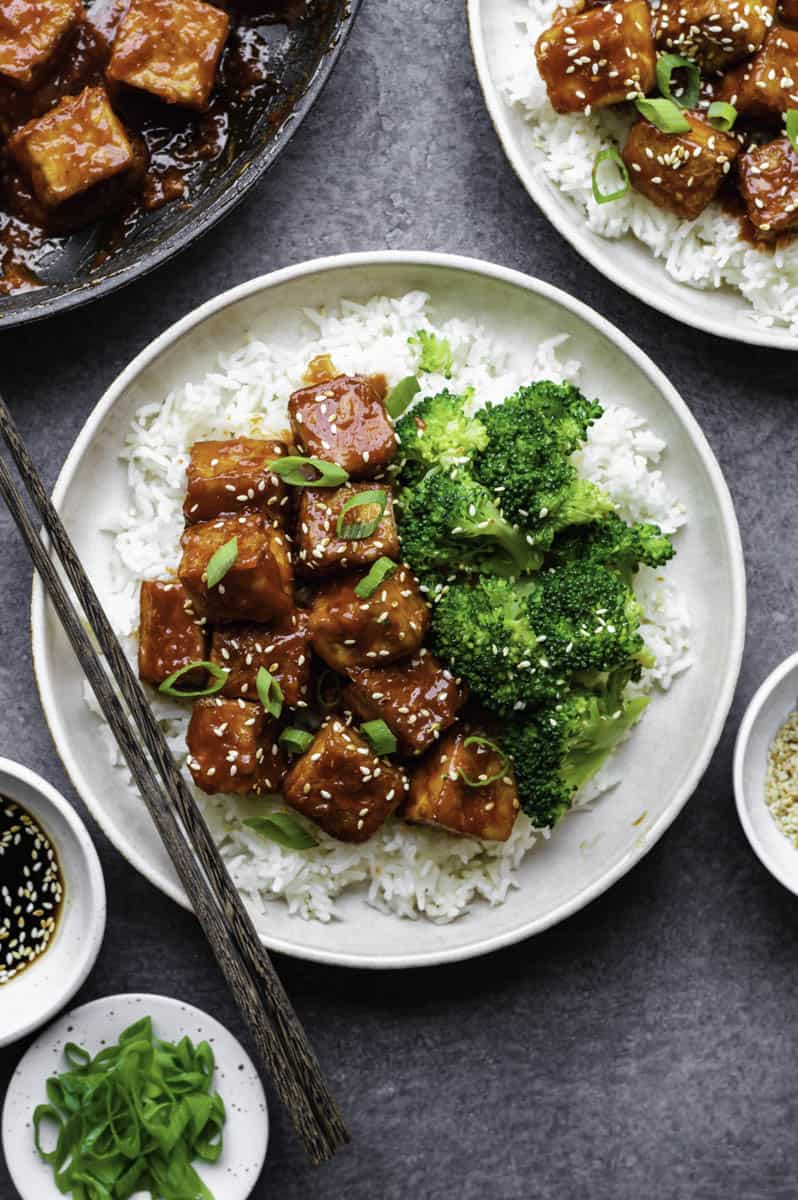 Gochujang tofu served with steamed broccoli over a bowl of white rice. Topped with sesame seeds and chopped green onion. Dark brown chopsticks placed on the left side of the bowl.