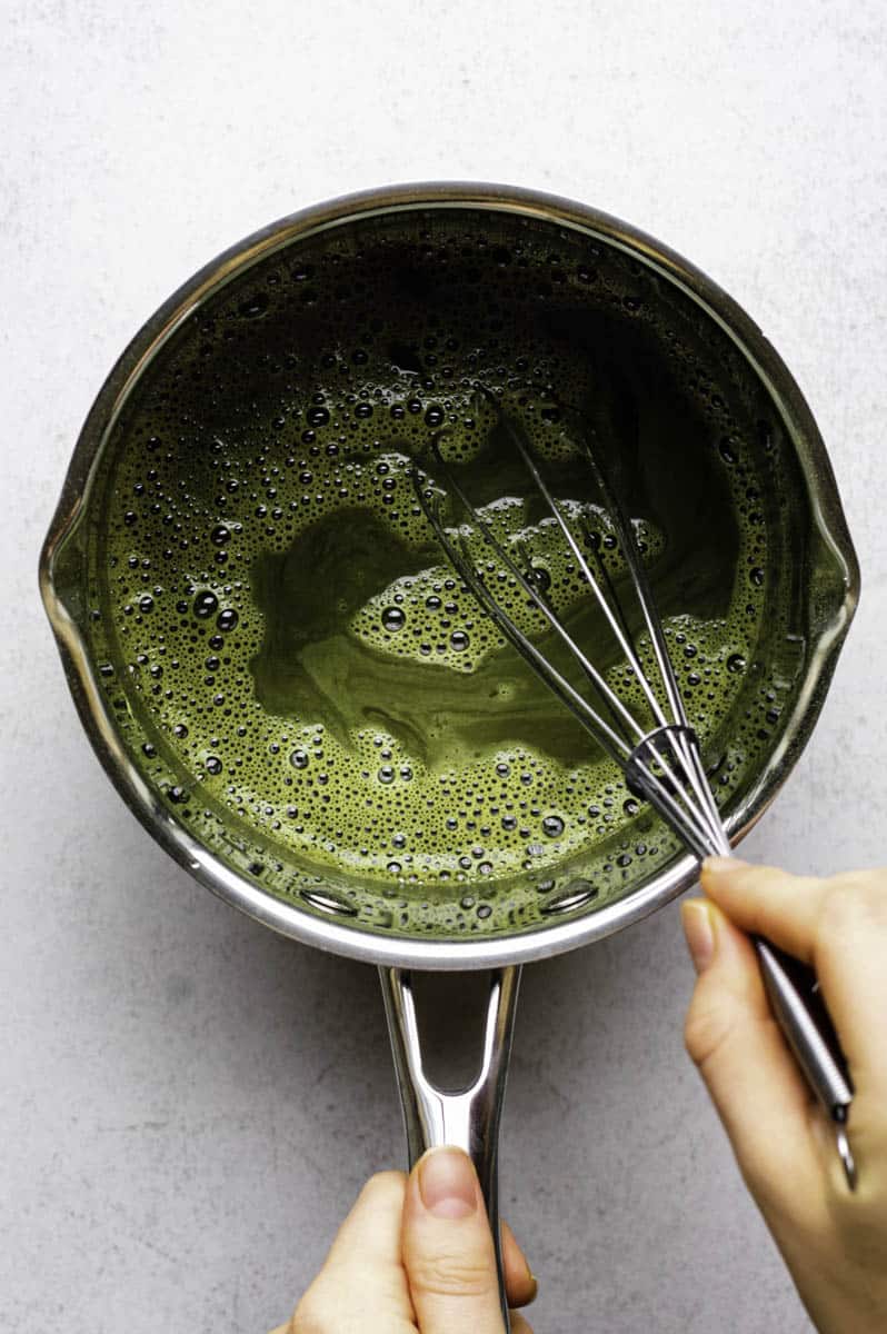 A hand mixing water, sugar, and matcha powder, in a small stainless-steel saucepan with a whisk.