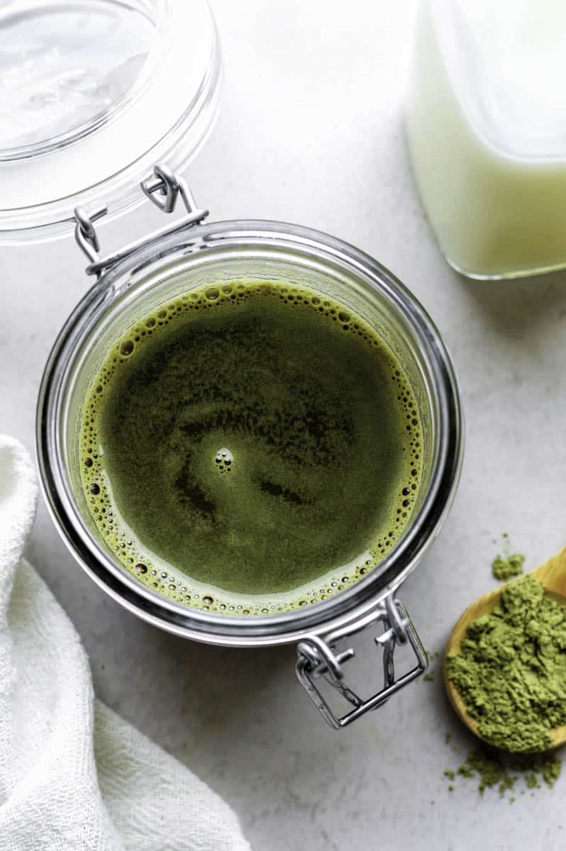 Matcha syrup in a glass jar. A white kitchen towel placed on one side of the jar, and a wooden spoon filled with matcha powder on the other.
