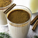 A glass of oat milk eggnog topped with ground cinnamon and a cinnamon stick.