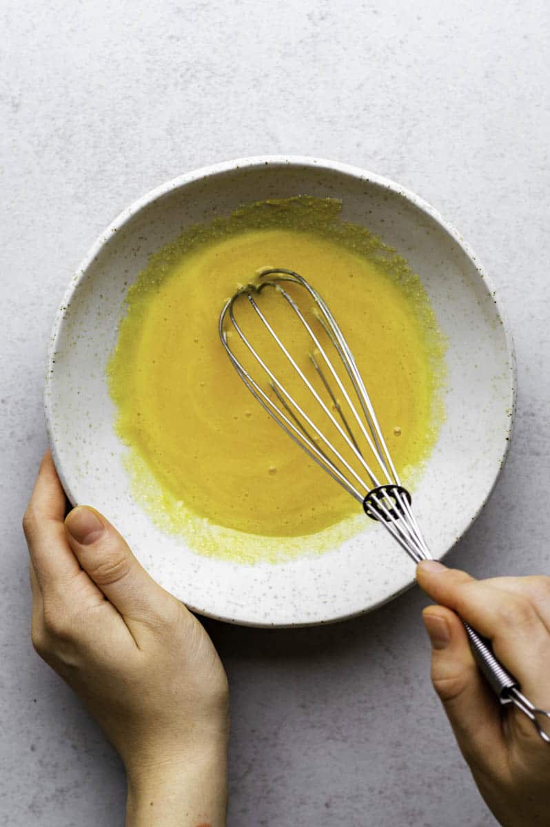 A white bowl filled with a mixture of egg yolks and sugar. A hand mixing it with a stainless-steel whisk.