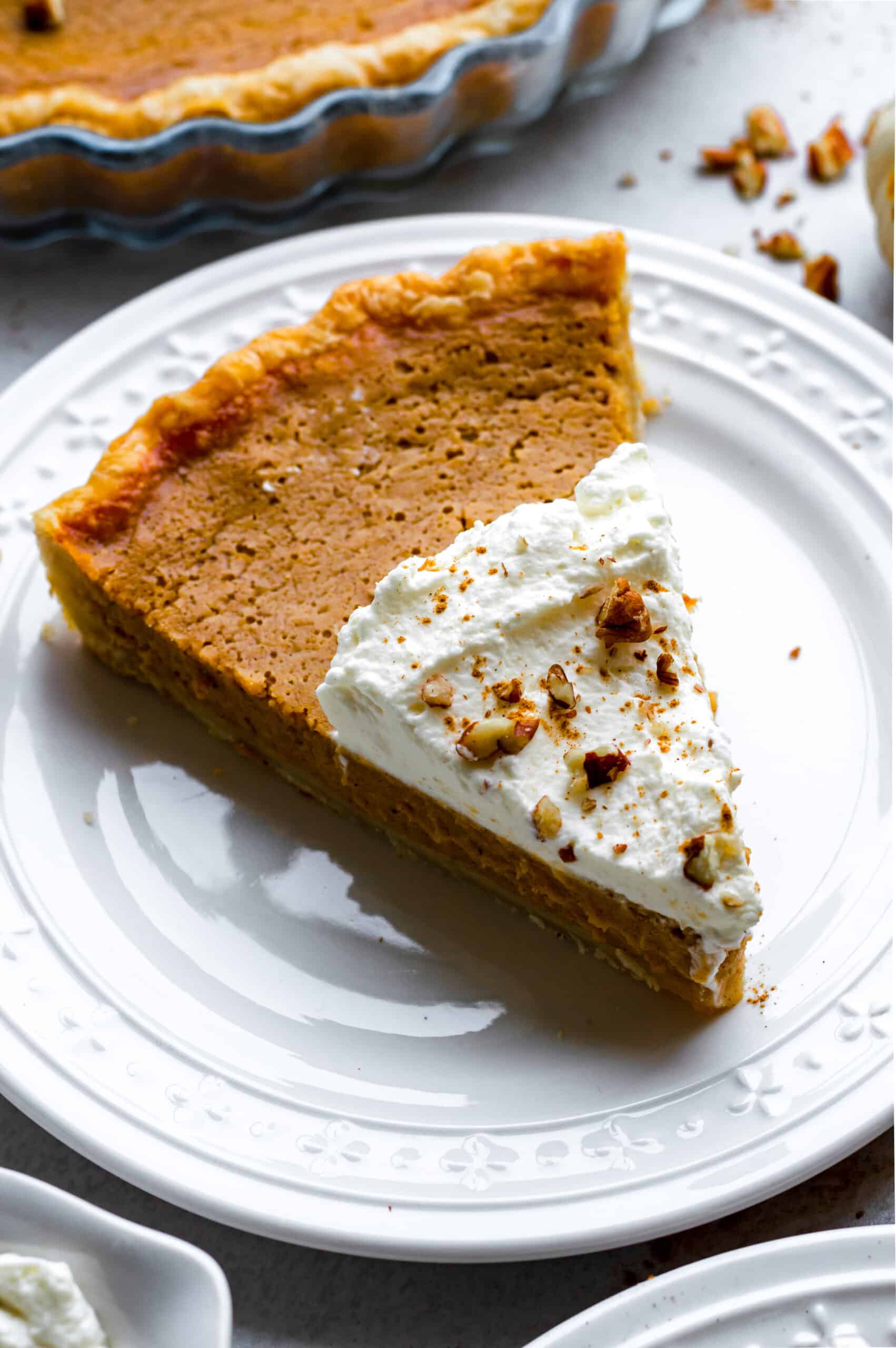 A piece of pumpkin pie without served on a white plate and topped with whipped cream and crushed pecans.