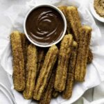 Vegan churros served in a white bowl layered with parchment paper with a small bowl full of hot chocolate in it.