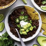 A white bowl filled with Beyond meat chili and topped with sour cream, cubed avocado, sliced jalapeño peppers, and cilantro.