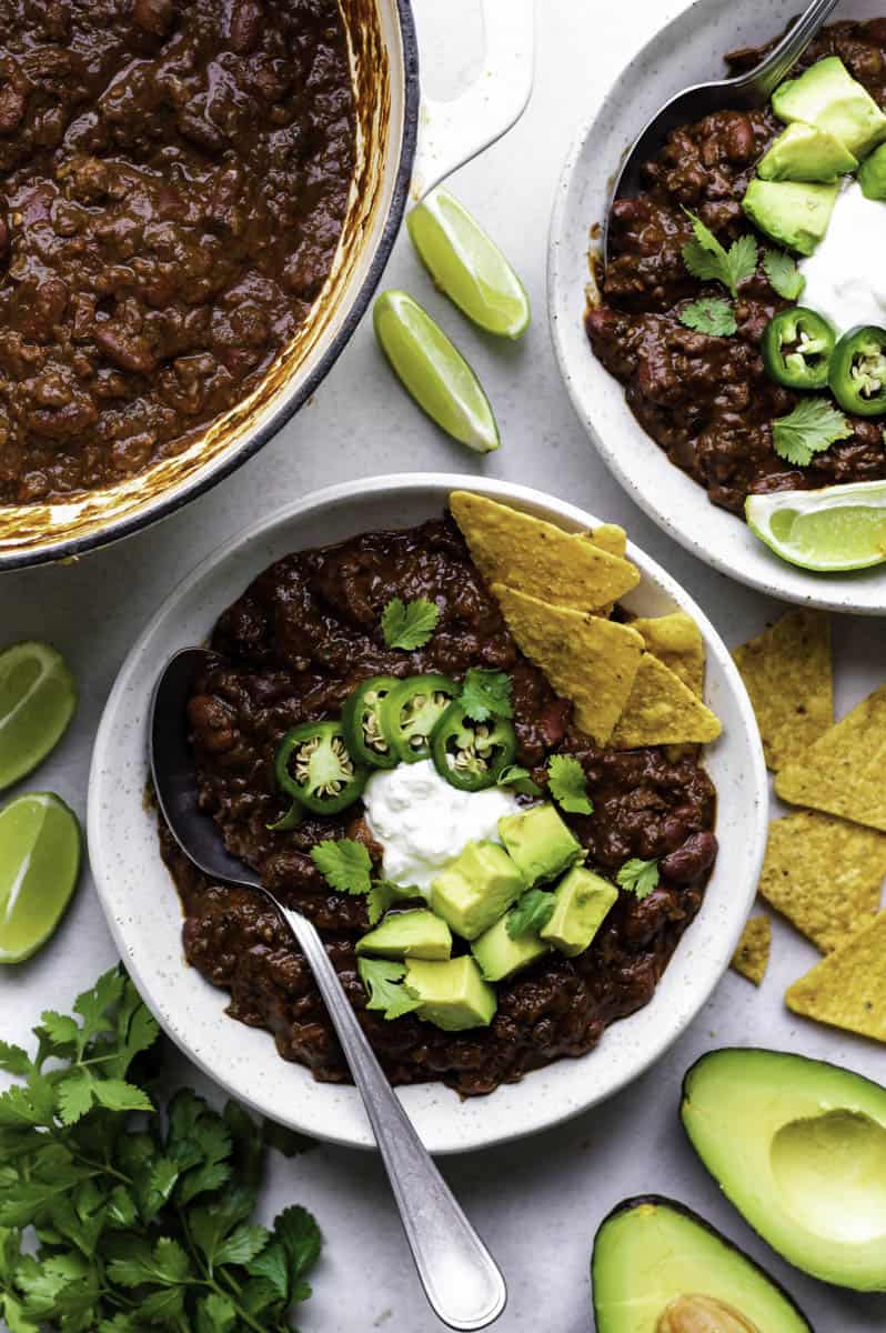 Two white bowls filled with Beyond meat chili and topped with sour cream, cubed avocado, sliced jalapeño peppers, and cilantro.