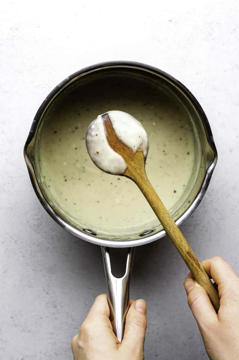 A saucepan filled with oat milk bechamel with a wooden spoon coated in sauce hovering over the pot.