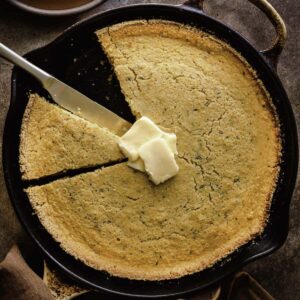 Vegan cornbread in a cast iron skillet with a slice taken out and vegan butter on top.