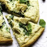 Vegan white pizza with spinach and artichokes, served on a piece of parchment paper, and topped with ground black pepper and basil leaves.