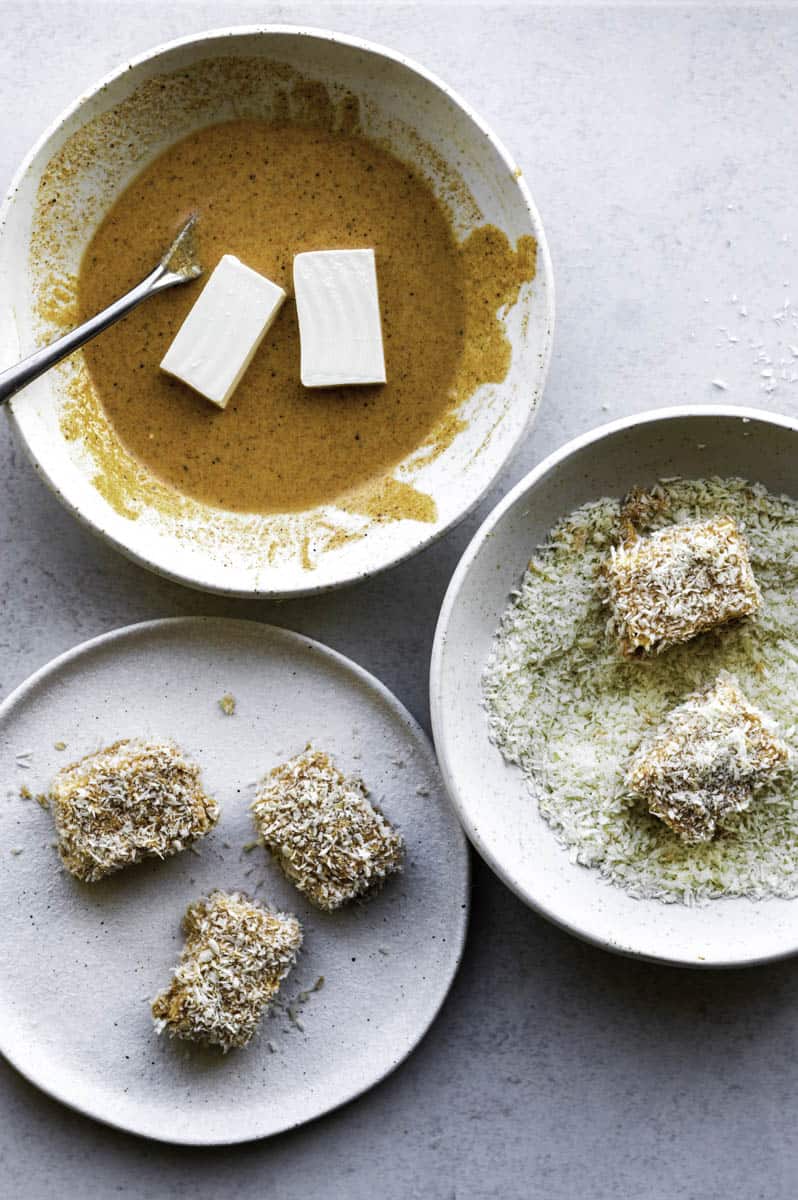 Three white bowl on a kitchen countertop. One is filled with wet batter, the second one is filled with breadcrumbs, and the third one with breaded tofu pieces.