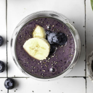 Blueberry banana smoothie in a glass with bananas and blueberries scattered around and chia seeds on top.