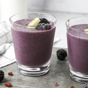 Two short glasses filled with purple blackberry banana smoothies and topped with frozen blackberries.