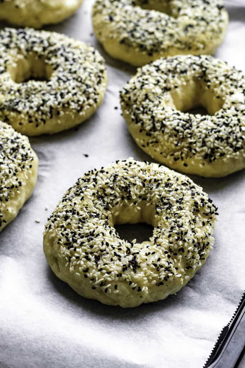 Unbaked bagels topped with everything bagel seasoing on a baking sheet.