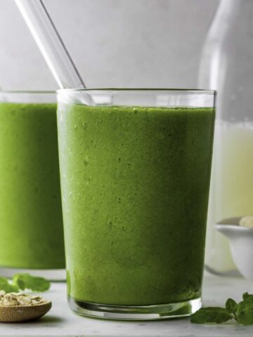 Two glasses filled with green smoothie with glass straw pointing out of them.