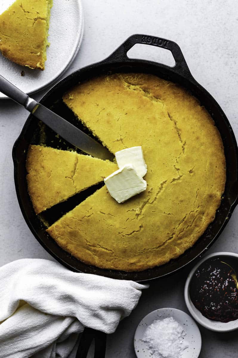 Cornbread in a cast-iron skillet with a slice take out, and topped with vegan butter.