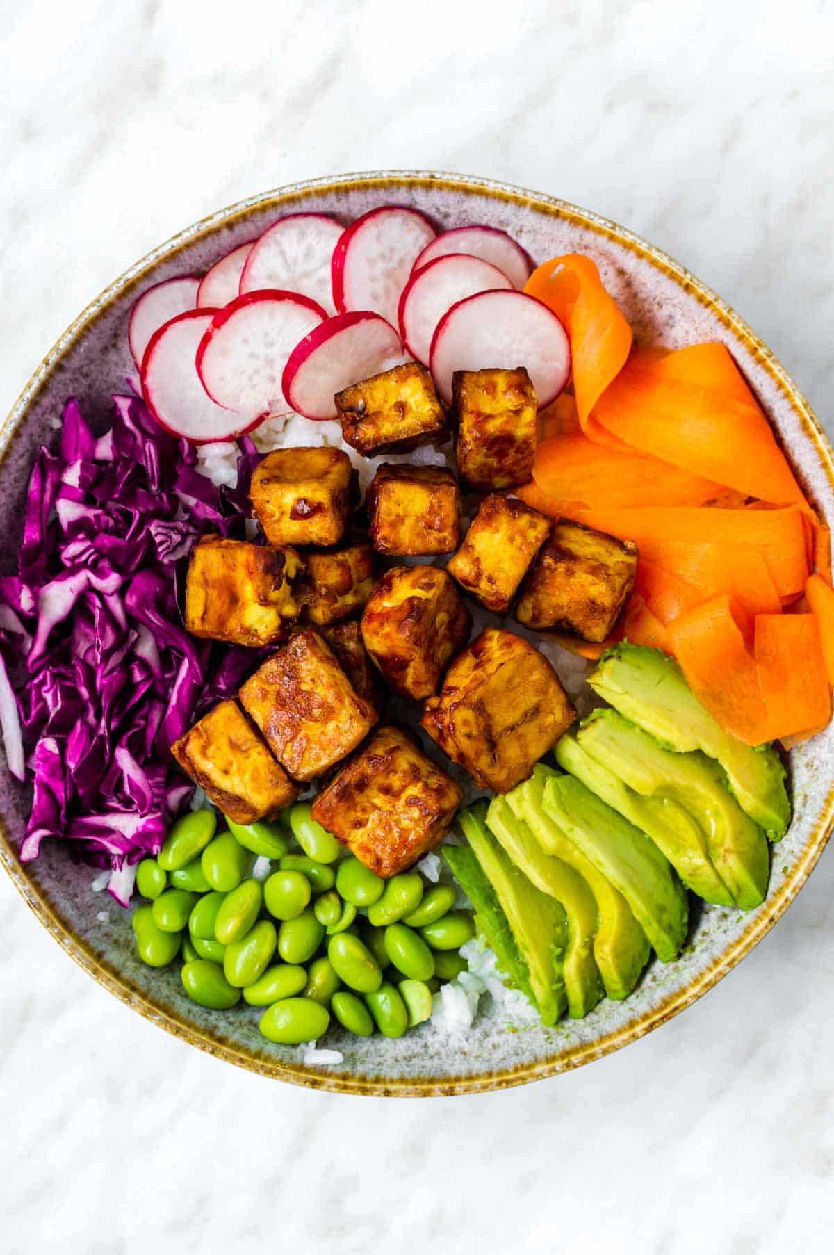 Assembled tofu buddha bowl with carrots, red cabbage, avocado, edamame and radishes.