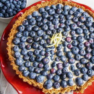 Red cake stand with a blueberry tart on top, garnished with lemon peel. Background of lemon and bowl of blueberries.