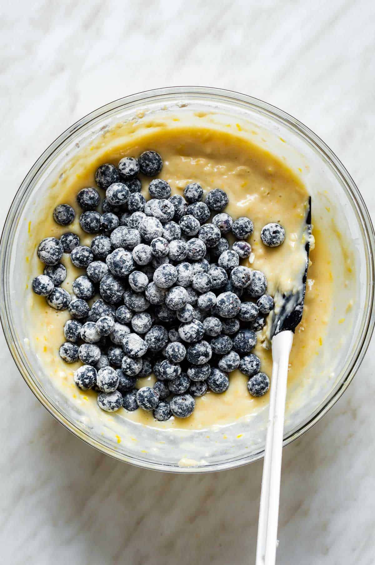 Adding blueberries to vegan muffin batter in a glass bowl.