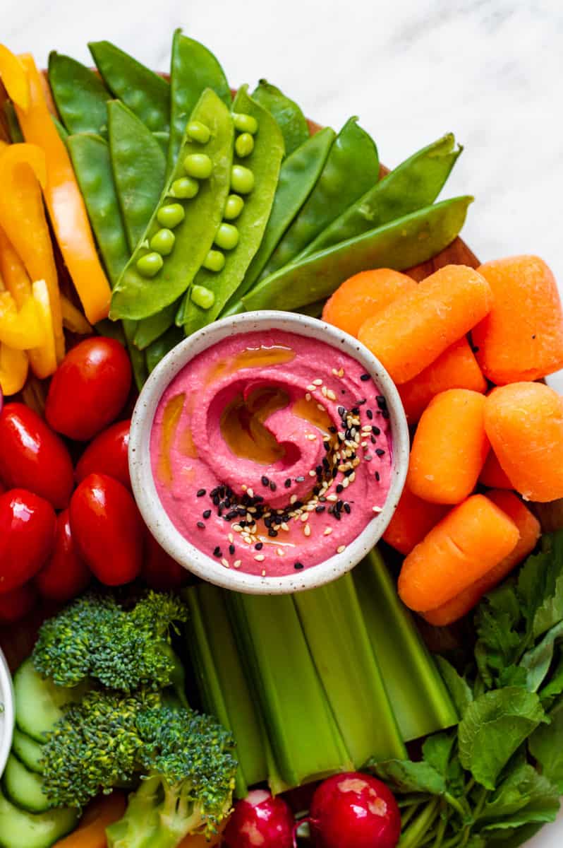 A small bowl filled with beet hummus. An array of vegetables surrounding the bowl.