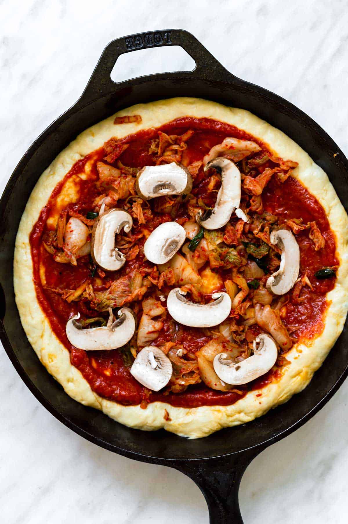 Pizza sauce with mushrooms and various toppings on Kimchi pizza dough on a cast-iron skillet.