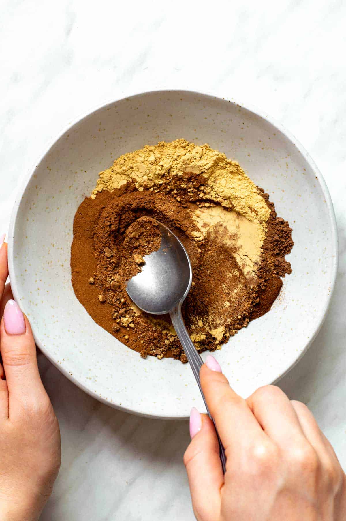 A hand mixing spices uisng a spoon in a white bowl.