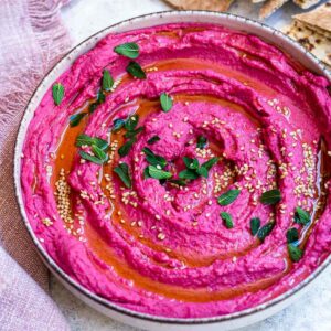 Beet hummus served in a bowl, topped with fresh mint leaves, sesame seeds, and olive oil.