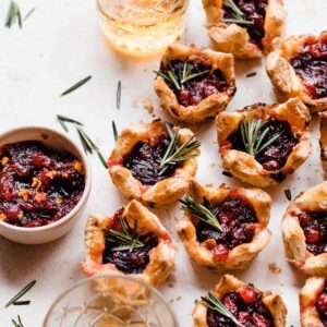 Puff pastry bites on a light surface surrounded by rosemary sprigs, glasses of rum, and a bowl of cranberry sauce.