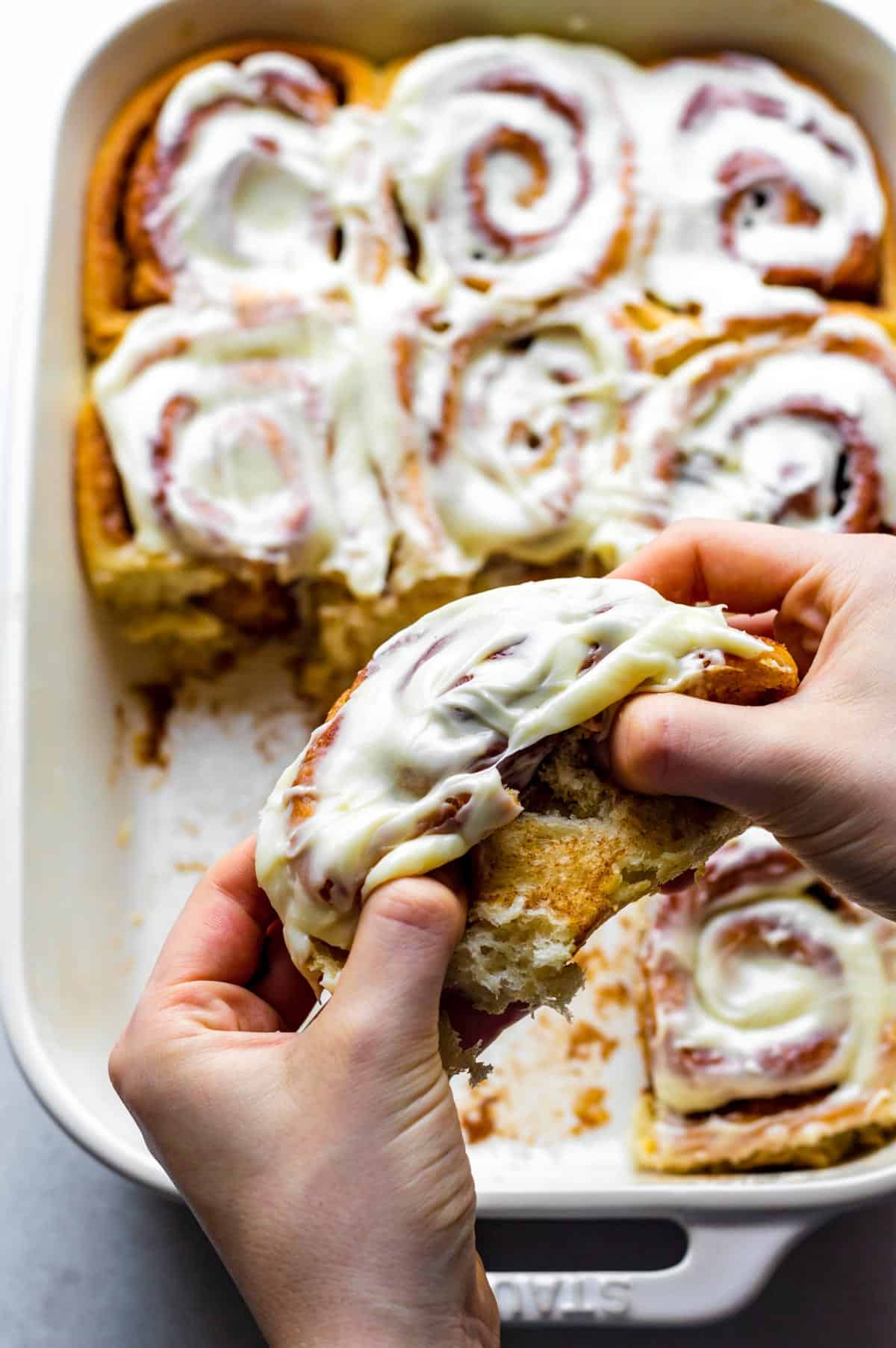 Two hands pulling apart a cinnamon roll topped with icing without cream cheese frosting.