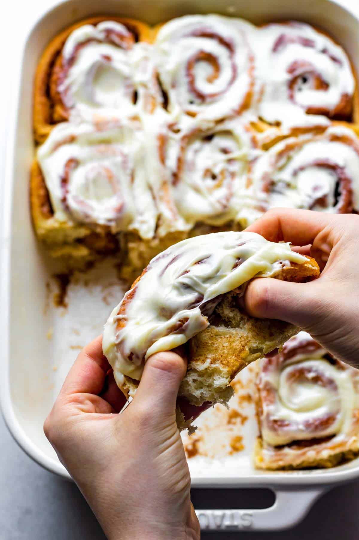 Two hands pulling apart a cinnamon roll topped with cream cheese frosting.