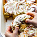 Two hands pulling apart a cinnamon roll topped with cream cheese frosting.