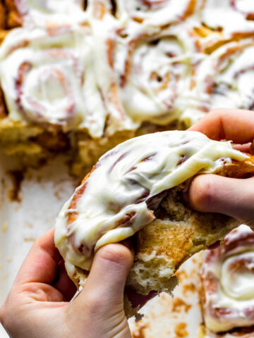 Two hands pulling apart a cinnamon roll topped with cream cheese frosting.