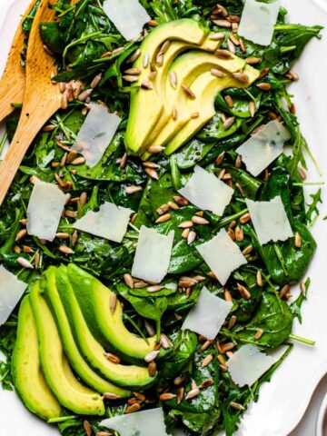 Spinach arugula salad served in a white platter and topped with avocado, parmesan and sunflower seeds.
