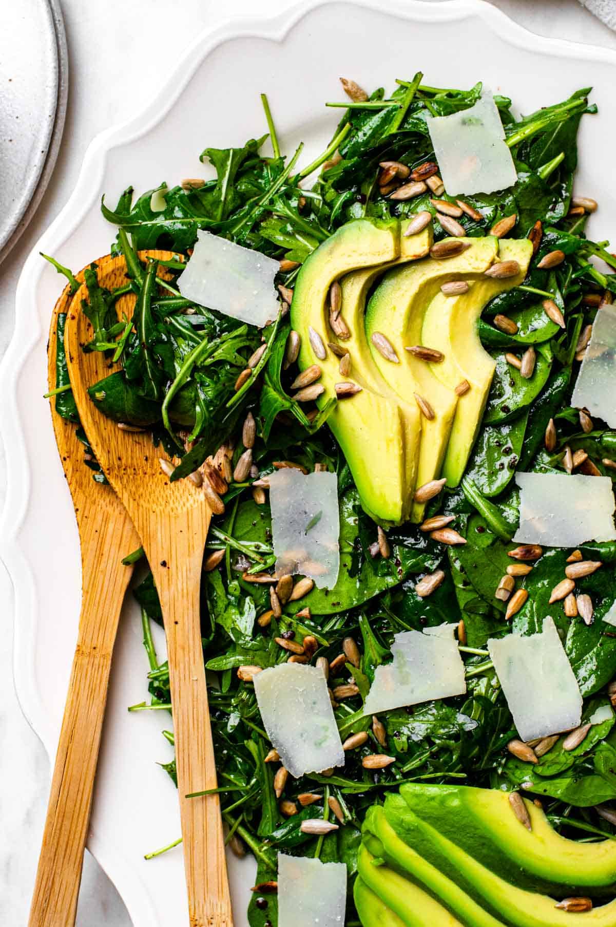 Green salad served on a white platter and topped with avocado, parmesan and sunflower seeds.