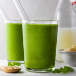 Two glasses filled with green smoothie with glass straw pointing out of them.