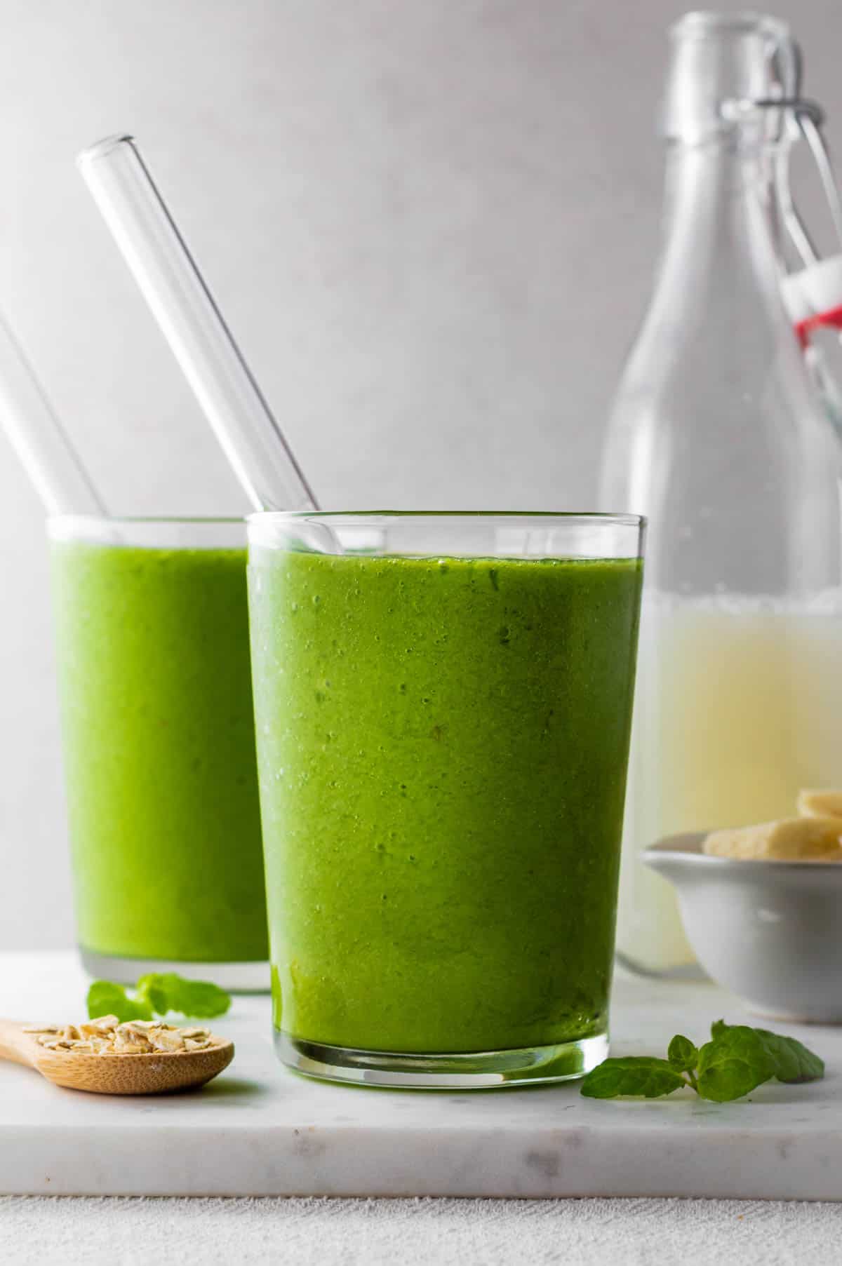 Two glasses filled with green smoothie with glass straw pointing out of them.