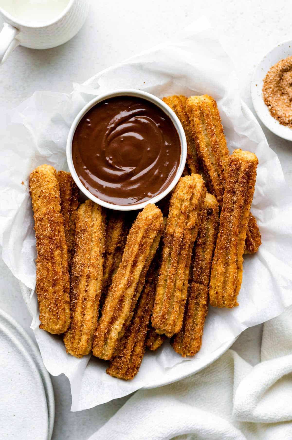 Vegan churros served in a white bowl layered with parchment paper with a small bowl full of hot chocolate in it.