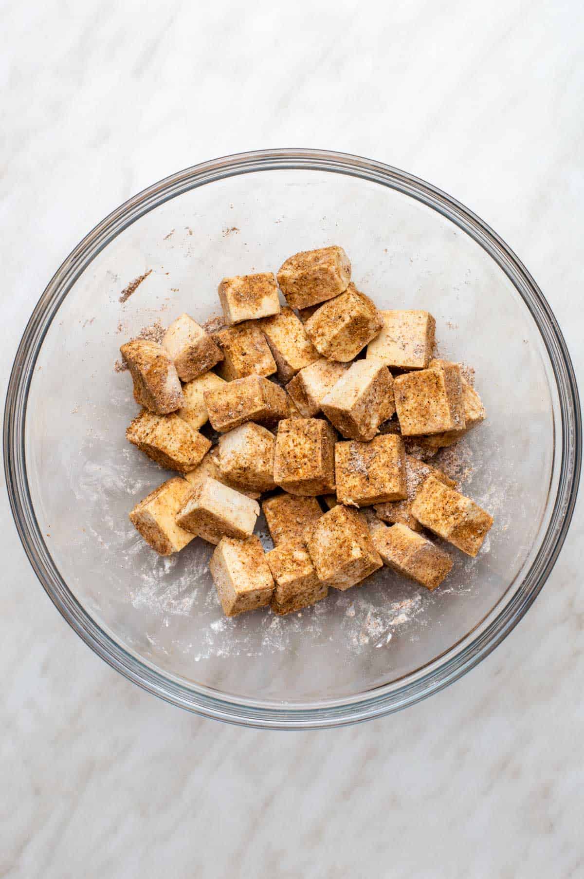 Tofu cubes tossed in spices and cornstarch in a glass mixing bowl.