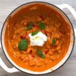 Romesco butter beans in a white Dutch oven topped with stracciatella cheese and basil leaves.