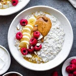 Overnight oats with water served in a white bowl and topped with banana, raspberries, peanut butter, and almonds.