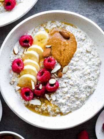 Overnight oats with water served in a white bowl and topped with banana, raspberries, peanut butter, and almonds.